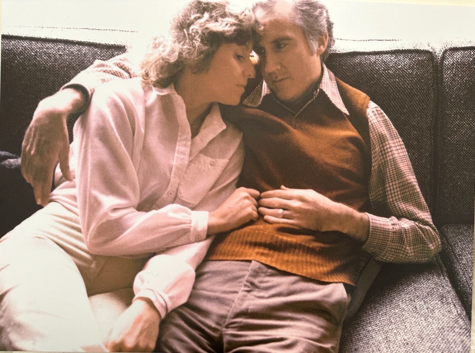 old color photo of Anne and Phil sitting on a grey couch, side by side, foreheads and fingers touching.