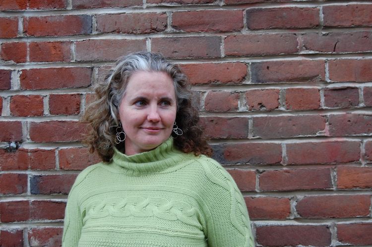 Christina, looking north, slightly smiling, with a brick wall behind her  