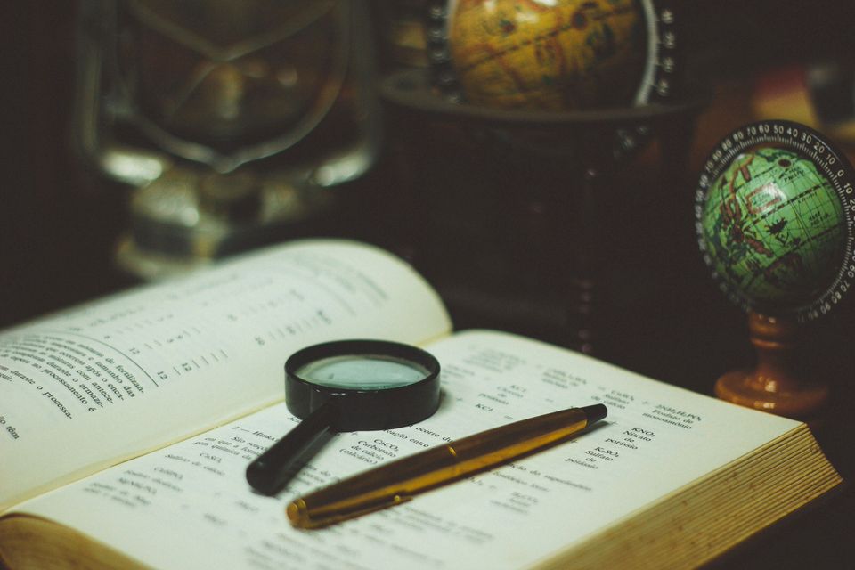 Open book with magnifying glass and pen on the page, on a desk with globes 